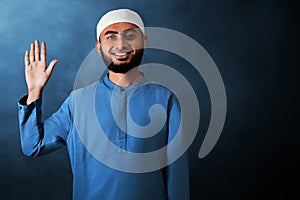 Portrait of handsome young asian muslim man with beard saying hello, waving hand gesture and smile