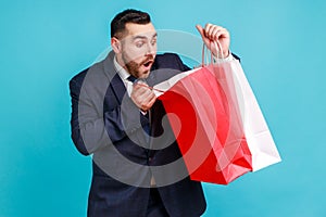 Portrait of handsome young bearded man in suit standing, holding red and white shopping bags and looking inside with shocked