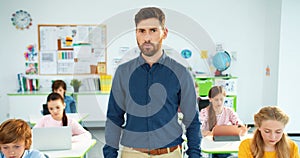 Portrait of handsome young Caucasian male tutor standing in classroom looking at camera and smiling. Children using