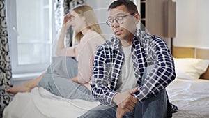 Portrait of handsome young Caucasian man in eyeglasses sitting on bed with blurred sad woman at background and talking