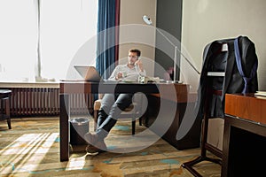 Portrait of handsome young entrepreneur speaking by phone and using laptop while working in comfortable hotel room or