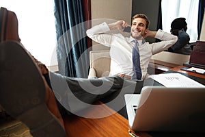 Portrait of handsome young entrepreneur speaking by phone and using laptop while working in comfortable hotel room or