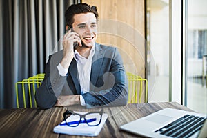 Portrait of handsome young male in glasses sitting at office desk with laptop computer and talking on mobile phone.