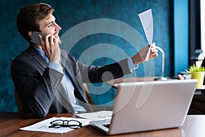 Portrait of handsome young male sitting at office desk with laptop computer and talking on mobile phone. Communication