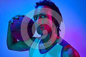 Portrait of handsome young man, basketball player posing with ball against gradient blue background in neon lights