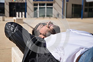 Portrait of handsome young man with beard, sunglasses, leather jacket and white shirt, lying on a stone bench. Concept beauty,