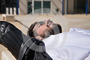 Portrait of handsome young man with beard, sunglasses, leather jacket and white shirt, lying on a stone bench. Concept beauty,