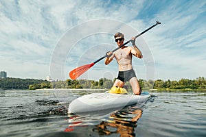Portrait of a handsome young man in casual clothes with an oar in his hand floating on the river on a sup board. Sport trains on a