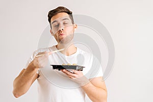 Portrait of handsome young man with enjoying eating fresh tasty sushi rolls with chopsticks on white isolated background