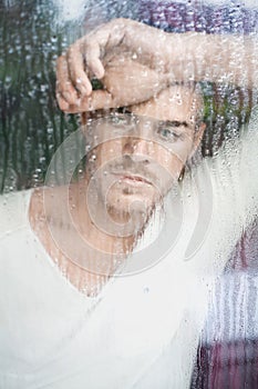 Portrait of handsome young man looking through wet windo