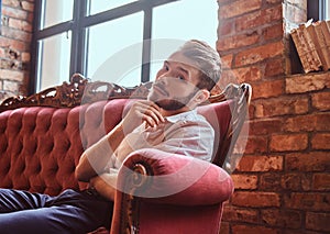 Portrait of a handsome young man with a stylish beard and hair elegantly dressed sitting on a red vintage sofa