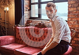 Portrait of a handsome young man with a stylish beard and hair elegantly dressed sitting on a red vintage sofa