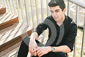 Portrait of a handsome young successful male brunette of a Turkish Mediterranean race sitting on the urban stairs in a black shirt