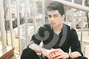 Portrait of a handsome young successful male brunette of a Turkish Mediterranean race sitting on the urban stairs in a black shirt