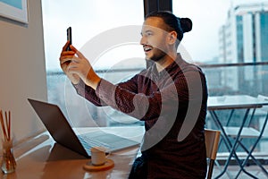 Portrait of happiness handsome man making selfie photo on smartphone, working at laptop, background of panoramic windows