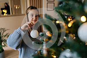 Portrait of happy adorable little girl enjoying process of decorating Christmas tree at home. Cute smiling child hanging