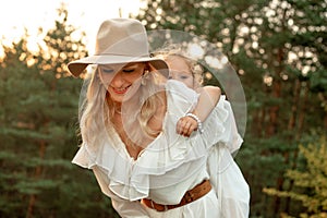 Portrait of happy adorable woman mother giving piggyback ride to little girl in forest illumined by sunset in summer.