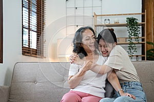 Portrait happy adult daughter and older mother hugging and holding hands, sitting on couch at home, young woman and