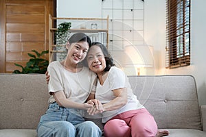 Portrait happy adult daughter and older mother hugging and holding hands, sitting on couch at home, young woman and