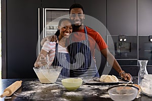 Portrait of happy african american couple in aprons preparing bread dough in kitchen