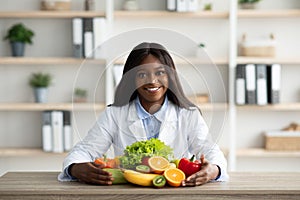 Portrait of happy african american dietitian in lab coat sitting with fresh fruits and vegetables on table