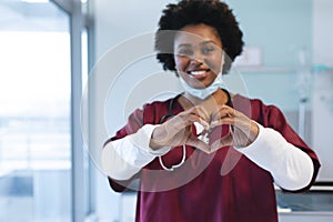 Portrait of happy african american female doctor wearing scrubs, forming hands in heart shape