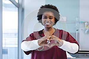 Portrait of happy african american female doctor wearing scrubs, forming hands in heart shape