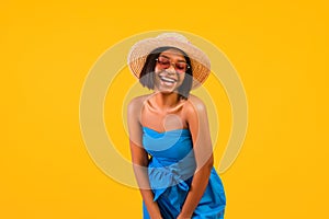 Portrait of happy African American lady in elegant dress, straw hat and sunglasses laughing on orange studio background