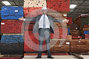 Portrait of happy African American male engineer standing with arms outstretched in front of stacked wooden planks