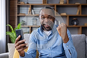 Portrait of a happy African American man sitting on the couch at home, using a mobile phone, and looking happy at the