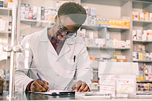 Portrait of a happy African American pharmacist writing prescription at workplace in modern pharmacy