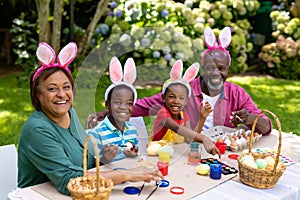 Portrait of happy african american siblings and grandparents in bunny ears painting easter eggs