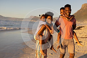 Portrait of happy african american young parents piggybacking son and daughter at beach against sky