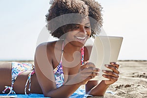Happy afro american woman lying on beach reading ebook