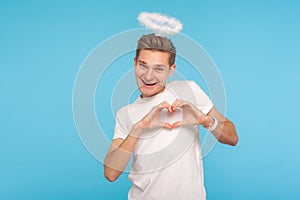 Portrait of happy angelic man with halo above head making heart shape with hands and smiling to camera