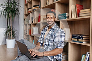 Portrait of happy arab man sitting with laptop and leaning on bookshelf, working on pc laptop and smiling at camera