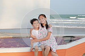 Portrait of happy Asian boy and girl sitting and hugging each other while sitting on sailboat and looking camera on beach at