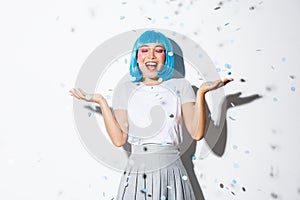 Portrait of happy asian girl in blue wig celebrating halloween, throwing confetti in the air, standing over white