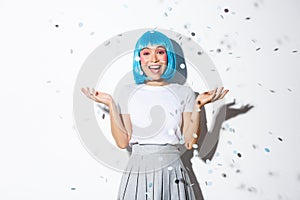 Portrait of happy asian girl in blue wig celebrating halloween, throwing confetti in the air, standing over white
