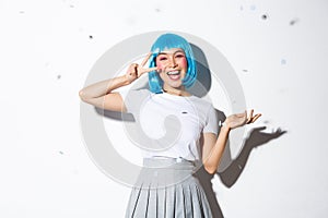 Portrait of happy asian girl in blue wig celebrating halloween, throwing confetti and showing kawaii peace gesture