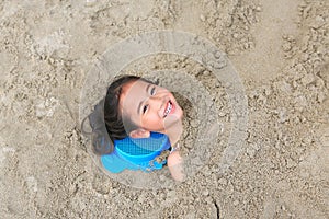 Portrait of happy asian little child girl buried in the sand at the beach. Close up kid playing with sand