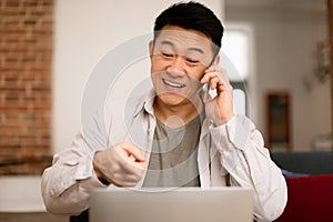 Portrait of happy asian man working from home, talking on cellphone and using laptop, having phone conversation