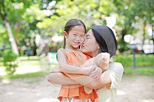 Portrait of happy Asian mother cuddle daughter and hugging teddy bear doll in the garden. Mom and child girl with love and