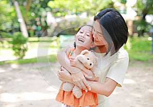 Portrait of happy Asian mother cuddle daughter and hugging teddy bear doll in the garden. Mom and child girl with love and