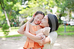 Portrait of happy Asian mother cuddle daughter and hugging teddy bear doll in the garden. Mom and child girl with love and