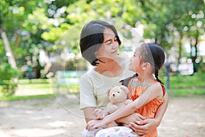 Portrait of happy Asian mother cuddle daughter and hugging teddy bear doll in the garden. Mom and child girl with love and