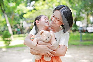 Portrait of happy Asian mother cuddle daughter and hugging teddy bear doll in the garden. Mom and child girl with love and