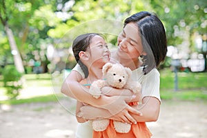 Portrait of happy Asian mother cuddle daughter and hugging teddy bear doll in the garden. Mom and child girl with love and