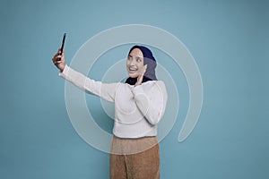 A portrait of a happy Asian Muslim woman wearing a white shirt and hijab, holding her phone, isolated by blue background