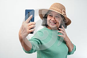 Portrait of happy Asian senior woman wearing sunglasses, a hat, and holding smartphone for a selfie, standing isolated on white